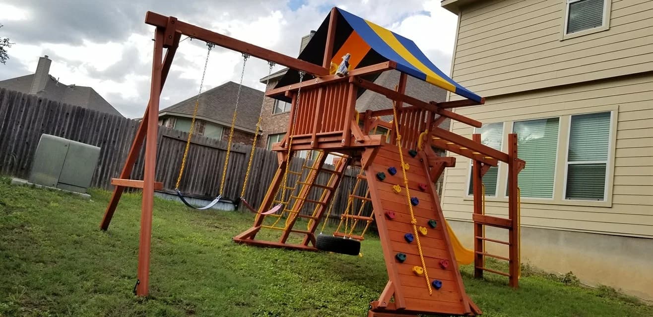 Wooden playset on a lawn between a house and a wooden fence