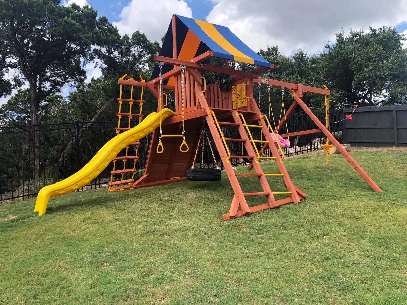 Wooden playset with a yellow slide and swings on a sloping lawn